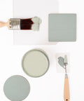 A paintbrush with greenish-grey paint, a round jar of matching paint, two painted swatches, and a palette knife with paint are arranged on a white background.