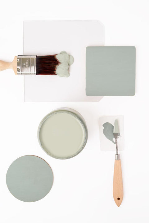 A paintbrush with greenish-grey paint, a round jar of matching paint, two painted swatches, and a palette knife with paint are arranged on a white background.