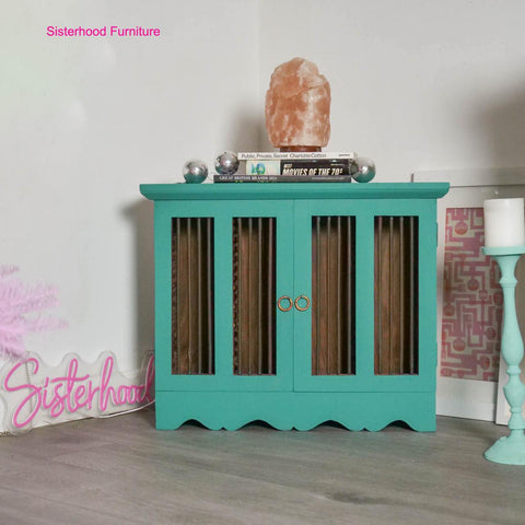A teal cabinet, featuring gold handles, stands on a wooden floor. It is topped with a large pink salt lamp and 3 small disco balls. A pink neon sign reading "Sisterhood" and a white candle are nearby.