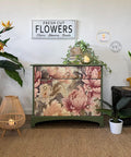 A cozy room corner featuring a green painted dresser with the Peaceful Peonies Decoupage Paper applied on the front. A sign reading "fresh cut flowers, stems - blooms - seeds," a small round table with plants, a lit lamp, and a potted plant on a mat.