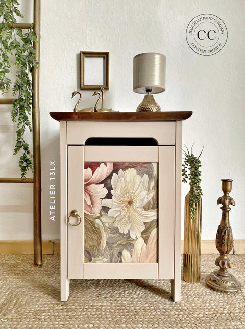 A small wooden cabinet with a floral design on the door sits on a woven rug. Atop the cabinet are two swan figurines and a small gold lamp. A golden frame hangs on the wall above. A wooden ladder with a plant and a gold candle holder are nearby.