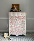 A vintage white dresser with pink floral patterns on its drawers stands against a gray wall. It has crystal knobs and a lit candle on top, along with a framed picture and decorative items. Books and a postcard are on the tiled floor beside it.