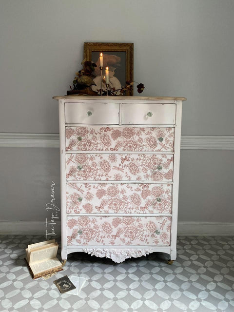 A vintage white dresser with pink floral patterns on its drawers stands against a gray wall. It has crystal knobs and a lit candle on top, along with a framed picture and decorative items. Books and a postcard are on the tiled floor beside it.