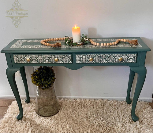 A blue-green vintage console table with intricate white filigree designs, gold knobs, and two drawers. A lit candle, beaded garland, and greenery decorate the top. The table sits on a fluffy rug, next to a glass jar with a plant.