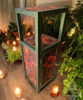 A small green wooden cabinet with red floral patterns on the door, shelf, and top, surrounded by lit candles and potted plants in a cozy, softly lit room.
