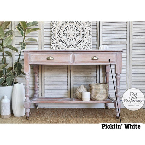 A beautifully restored wooden console table with two drawers, featuring ornate decorations and baskets on the shelf below. It stands against a decorative wall panel with a round, intricate art piece. Potted plants and vases add a natural touch.