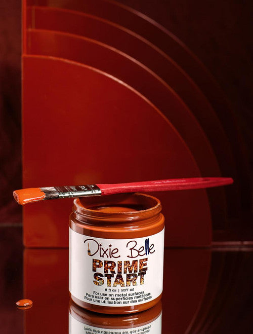 A jar of Dixie Belle Prime Start paint with an orange paint-dipped brush resting on top. Some paint drips down the jar, and the background features layered, curved, reddish-brown panels.