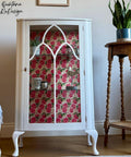 White cabinet with floral pink and red decoupage paper, displaying various items inside. It stands next to a wooden plant stand holding a green plant. The scene is set against a wooden floor and light-colored wall with decorative text.