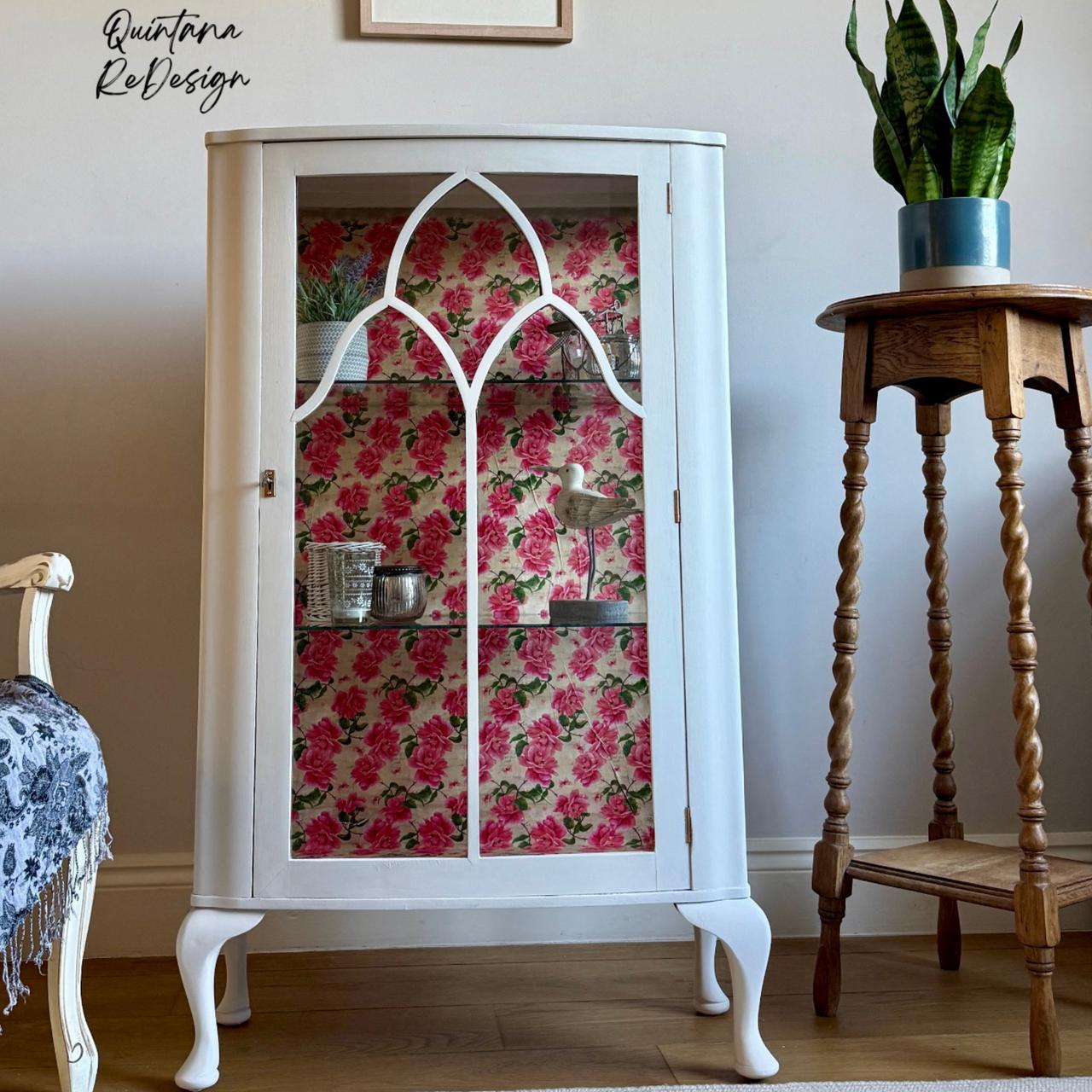 White cabinet with floral pink and red decoupage paper, displaying various items inside. It stands next to a wooden plant stand holding a green plant. The scene is set against a wooden floor and light-colored wall with decorative text.