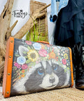 A vintage suitcase with an orange border features a painted raccoon wearing a colorful flower crown. The suitcase is positioned on a woven surface with a rustic wood backdrop, green plants, cowboy boots, and a denim jacket draped nearby. The watermark logo "Tracey's Fancy" is visible in the background.