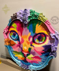 A colorful, abstract portrait of a cat fills an ornate, multicolored oval frame. The cat's face is depicted with vibrant hues of pink, orange, green, and blue. The frame features intricate floral designs and flourishes, adding elegance to the vivid artwork.