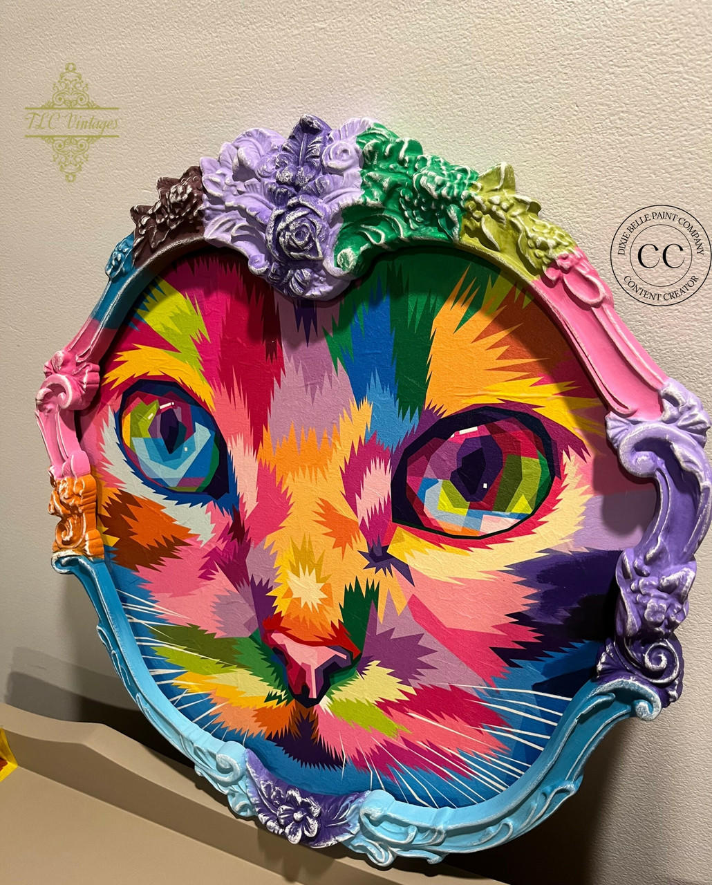 A colorful, abstract portrait of a cat fills an ornate, multicolored oval frame. The cat's face is depicted with vibrant hues of pink, orange, green, and blue. The frame features intricate floral designs and flourishes, adding elegance to the vivid artwork.