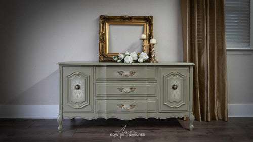 A vintage light green dresser with ornate gold handles sits against a wall. On top are a gold picture frame, a bouquet of white flowers, and two gold candlesticks. Gold curtains hang beside a window with blinds.