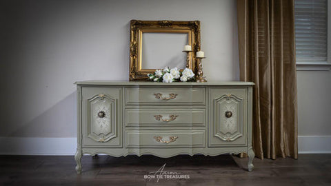 A vintage light green dresser with ornate gold handles sits against a wall. On top are a gold picture frame, a bouquet of white flowers, and two gold candlesticks. Gold curtains hang beside a window with blinds.