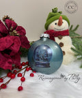 A painted Christmas ornament with a snowy winter scene sits on a white fur surface next to red flowers, berries, and a snowman decoration. Text reads “Reimagined by Patty” and a logo is in the top right corner.