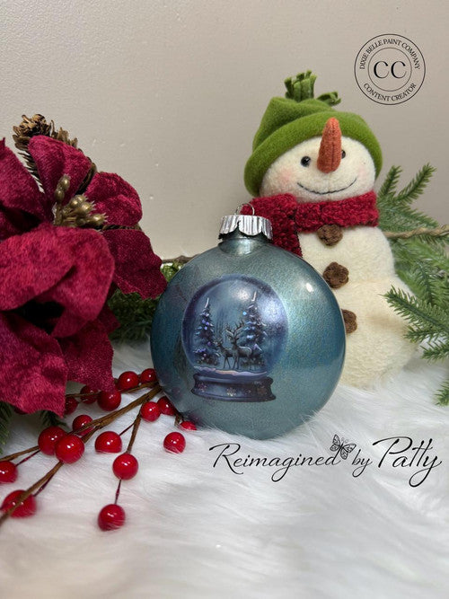 A painted Christmas ornament with a snowy winter scene sits on a white fur surface next to red flowers, berries, and a snowman decoration. Text reads “Reimagined by Patty” and a logo is in the top right corner.