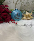 A blue Christmas ornament with a snowman design sits on white faux fur, surrounded by red poinsettias, gold flowers, greenery, and a glittery "Merry Christmas" sign in the background.