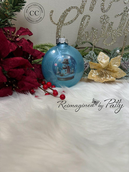A blue Christmas ornament with a snowman design sits on white faux fur, surrounded by red poinsettias, gold flowers, greenery, and a glittery "Merry Christmas" sign in the background.