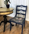 A vintage wooden chair painted in navy blue with a cushion featuring a colorful rooster and floral design sits beside a round, antique dark wooden table. A potted plant with variegated leaves rests on the table, set against a white paneled wall.