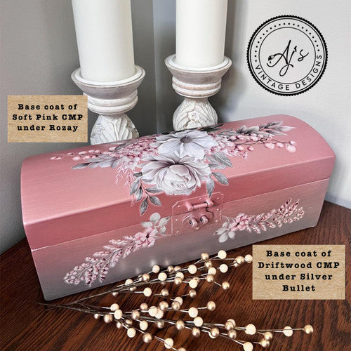 A pink and silver decorative box with white and pink flowers sits on a table. Two white candles in ornate holders are behind it. Text notes the box’s base coats: “Soft Pink CMP under Rozay” and “Driftwood CMP under Silver Bullet.”.