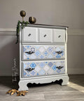 A gray 3 drawer dresser. Sapphire Sonata Transfer is seen applied to the bottom two drawers, the bottom two drawers have long intricate hardware, the top drawer has knobs hardware. Staged with bronze cups with a vine  on top of the piece and large gold leaves on the floor.