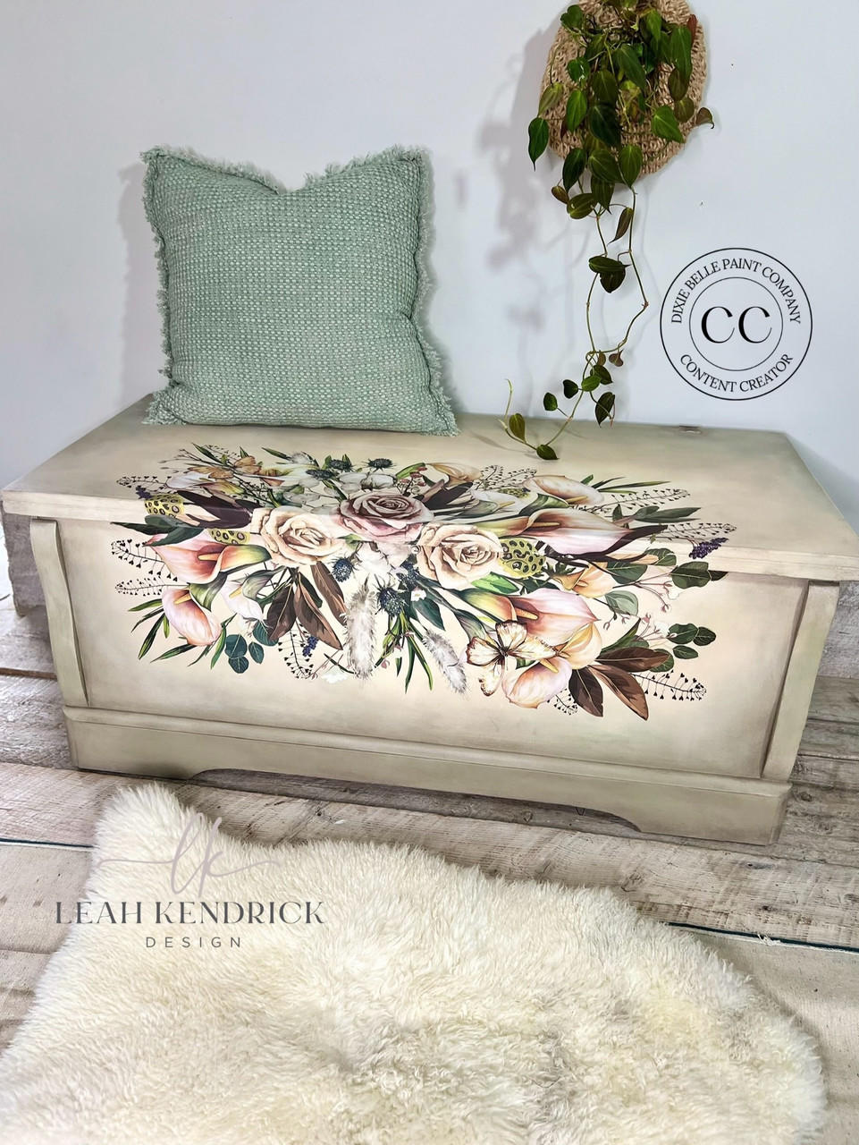 A wooden chest, decorated with an intricate floral design featuring roses and foliage, is placed against a white wall. A green textured pillow rests on top of the chest. A fluffy rug lies in front.