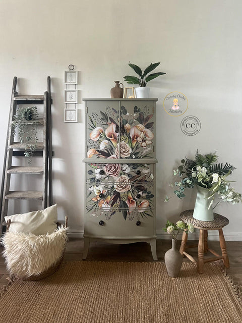 A well-decorated room features a hand-painted cabinet with floral designs. To the left of the cabinet, there is a wooden ladder adorned with greenery, and a wicker basket with a fluffy blanket and pillow. To the right, there are plants on a stool and in a vase.