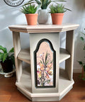 A vintage-style hexagonal side table with three shelves. The front panel is decorated with a butterfly and floral design. The tabletop is adorned with three potted plants. The background includes additional green plants. A logo reading "AJ's Vintage Designs" is visible.
