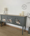 A vintage-style sideboard painted in dark grey with floral designs on its doors. It is positioned against a plain white wall. The sideboard is decorated with a lamp and two candles.