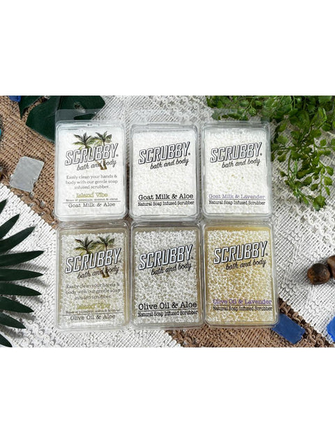 Image of Scrubby Soap Bath & Body group. Island Vibe Goat Milk & Aloe, Goat Milk & Aloe, Goat Milk & Lavender, Island Vibe Olive Oil & Aloe, Olive Oil & Aloe, Olive Oil & Lavender. Surrounded by faux plants and leaves, blue sea glass, and brown pebbles.