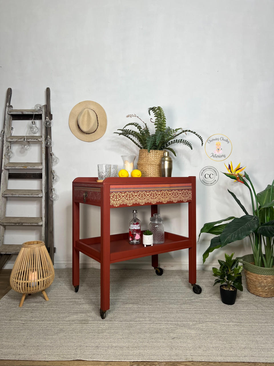 Red painted drink cart with black wheels. Staged with glasses, lemons, a shaker cup, a candle inside a vase and a wicker basket containing faux greenery on top.