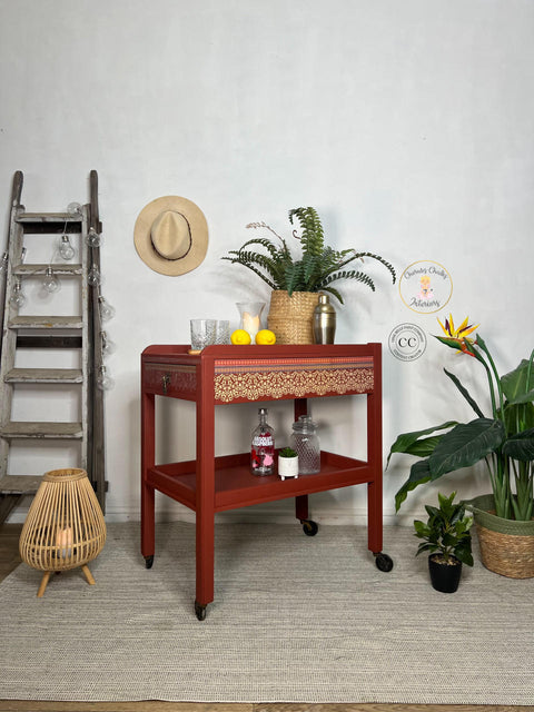 Red painted drink cart with black wheels. Staged with glasses, lemons, a shaker cup, a candle inside a vase and a wicker basket containing faux greenery on top.