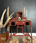 Red painted desk transformation with wooden spindles. Staged with an open book and two lit candlesticks on top. Large pieces of driftwood is placed next to the piece along with some wooden shoe inserts on the floor.