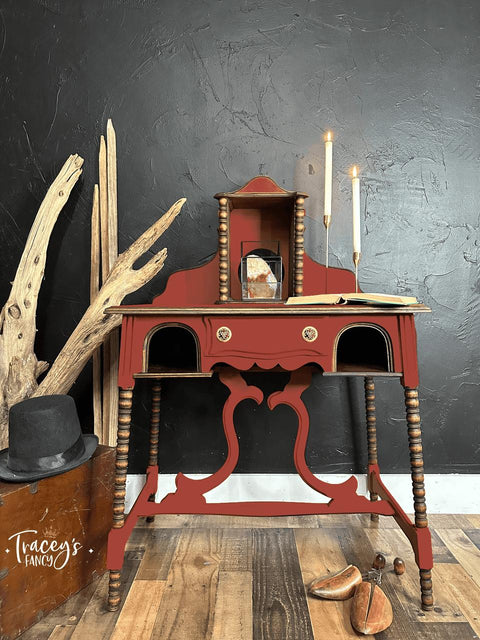 Red painted desk transformation with wooden spindles. Staged with an open book and two lit candlesticks on top. Large pieces of driftwood is placed next to the piece along with some wooden shoe inserts on the floor.