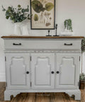 Baja Gray Silk All-In-One paint is perfect for painting kitchen cabinets, bathroom cabinets and painting furniture. Baja Gray Silk All-In-One Mineral Paint on small cabinet. The cabinet has black hardware and a natural wood top. The piece is staged with candle sticks and vintage flower arrangements against a white wall with a painting of various vegetation.