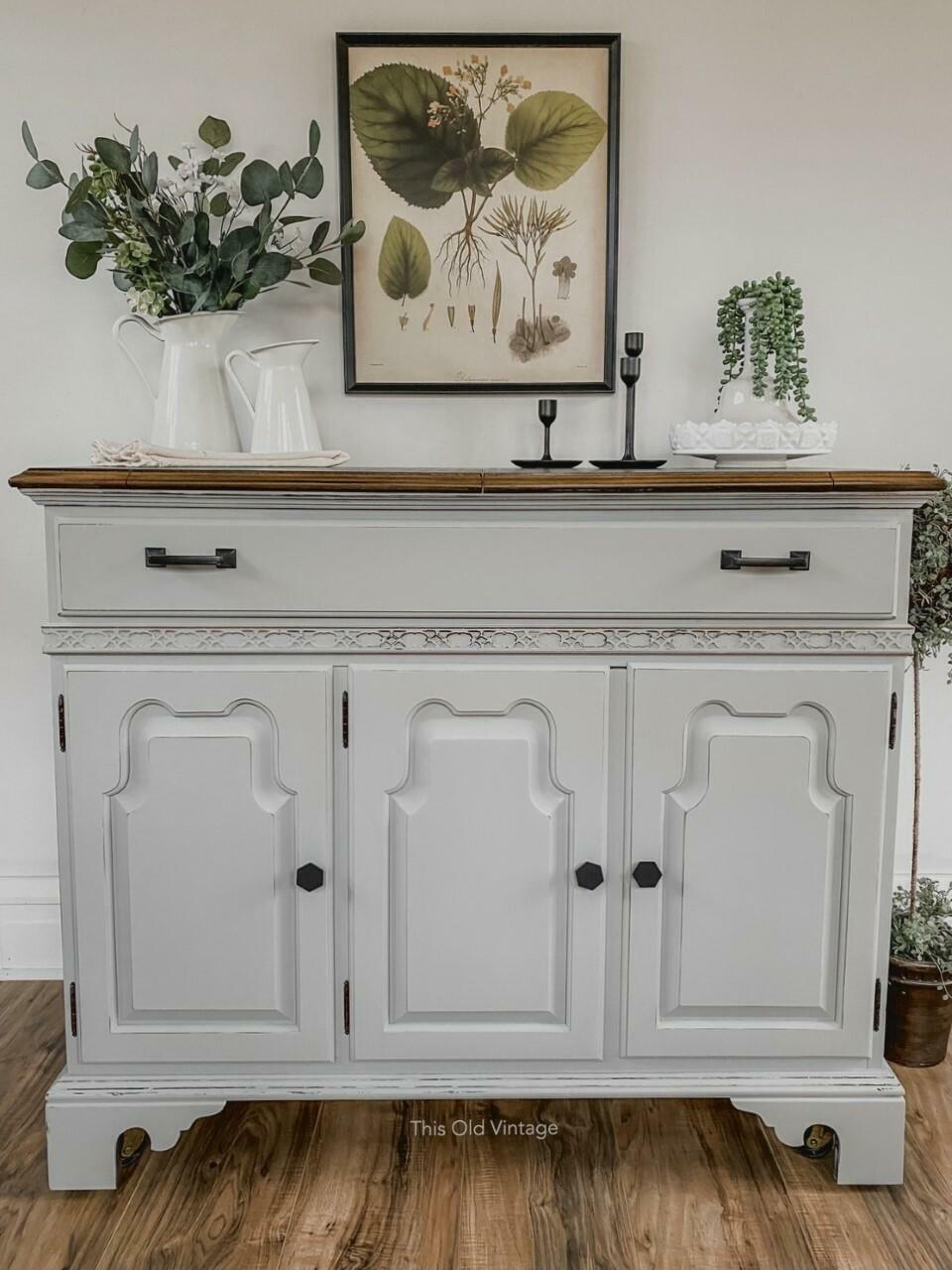 Baja Gray Silk All-In-One paint is perfect for painting kitchen cabinets, bathroom cabinets and painting furniture. Baja Gray Silk All-In-One Mineral Paint on small cabinet. The cabinet has black hardware and a natural wood top. The piece is staged with candle sticks and vintage flower arrangements against a white wall with a painting of various vegetation.
