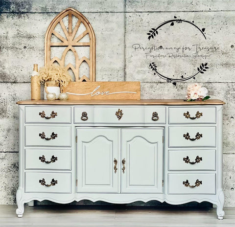 Baja Gray Silk All-In-One paint is perfect for painting kitchen cabinets, bathroom cabinets and painting furniture. Baja Gray Silk All-In-One Mineral Paint on a cabinet with bronze hardware. The piece is staged with a sign that says "Love" and a flower arrangement. The cabinets sits in front of a white brick wall.