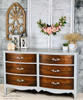 Baja Gray Silk All-In-One paint is perfect for painting kitchen cabinets, bathroom cabinets and painting furniture. The image shows Baja Gray Silk All-In-One paint on a waist high dresser. The dresser is painted with while the drawers are a beautiful natural wood. the piece is staged with flowers against a brick wall.