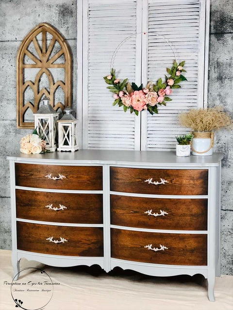 Baja Gray Silk All-In-One paint is perfect for painting kitchen cabinets, bathroom cabinets and painting furniture. The image shows Baja Gray Silk All-In-One paint on a waist high dresser. The dresser is painted with while the drawers are a beautiful natural wood. the piece is staged with flowers against a brick wall.