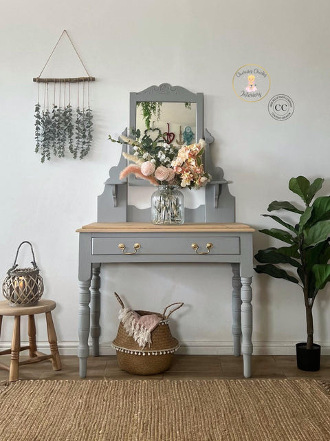 Baja Gray Silk All-In-One paint is perfect for painting kitchen cabinets, bathroom cabinets and painting furniture. This image shows Baja Gray Silk All-In-One Mineral Paint on a one drawer vanity with gold hardware and a natural wood top. There is a small stool next to the vanity along with a plant and a container holding a blanket. A flower arrangement sits atop the vanity to complete the piece.