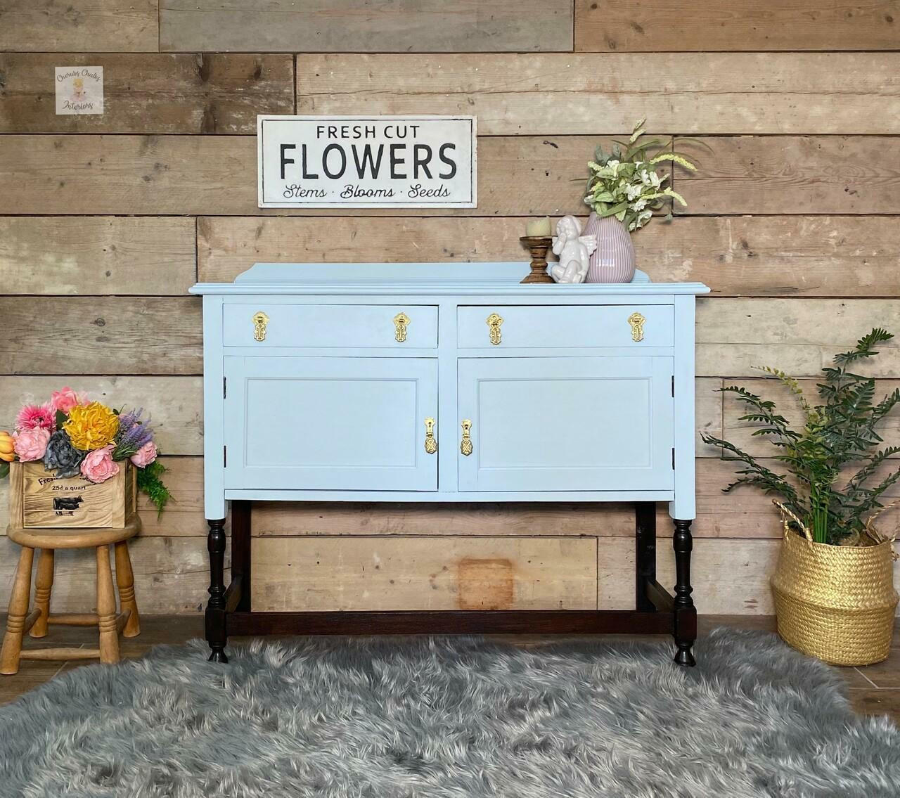 Bay Breeze Silk All-In-One paint is perfect for painting kitchen cabinets, bathroom cabinets and painting furniture.
Beautiful entrance table painted in Bay Breeze Silk Paint with gold hardware staged in front of a rustic wall and on top gray fur carpet. Beside piece is a gold woven basket,  and wooden stool with wooden box and multicolor flowers.