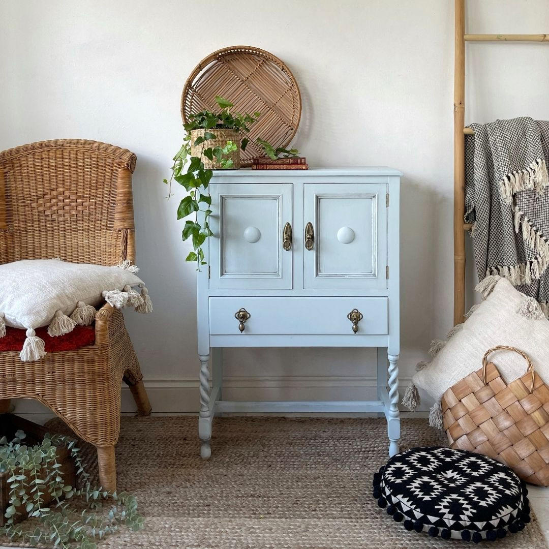 Silk All-In-One Mineral Paint Bay Breeze Silk Paint  is perfect for painting kitchen cabinets, bathroom cabinets and painting furniture.
Image shows a cabinet painted in Bay Breeze Silk Paint with some Black Glaze on the door panels and corners. Staged on a rattan-type rug with boho style decor around piece, staged on top of furniture is woven planet and woven tray place besides each other.