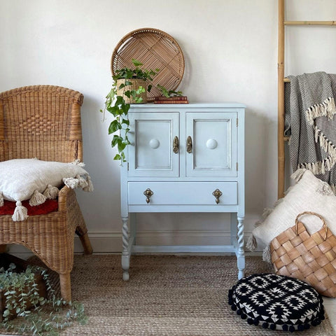 Silk All-In-One Mineral Paint Bay Breeze Silk Paint  is perfect for painting kitchen cabinets, bathroom cabinets and painting furniture.
Image shows a cabinet painted in Bay Breeze Silk Paint with some Black Glaze on the door panels and corners. Staged on a rattan-type rug with boho style decor around piece, staged on top of furniture is woven planet and woven tray place besides each other.