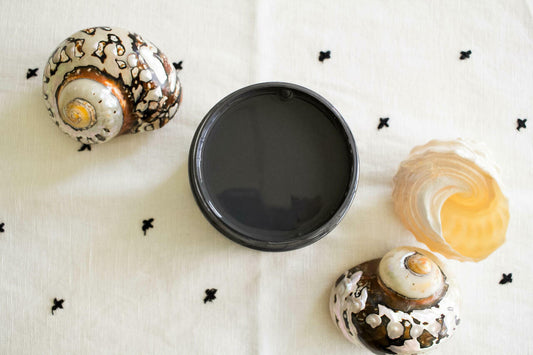 Black Sands Silk All-In-One paint is perfect for painting kitchen cabinets, bathroom cabinets and painting furniture. Open Jar of Black Sand Silk All in one mineral paint staged on a white cloth with black X's. White and gold conch shells on either side of open jar with white and orange seashell next to conch shell.