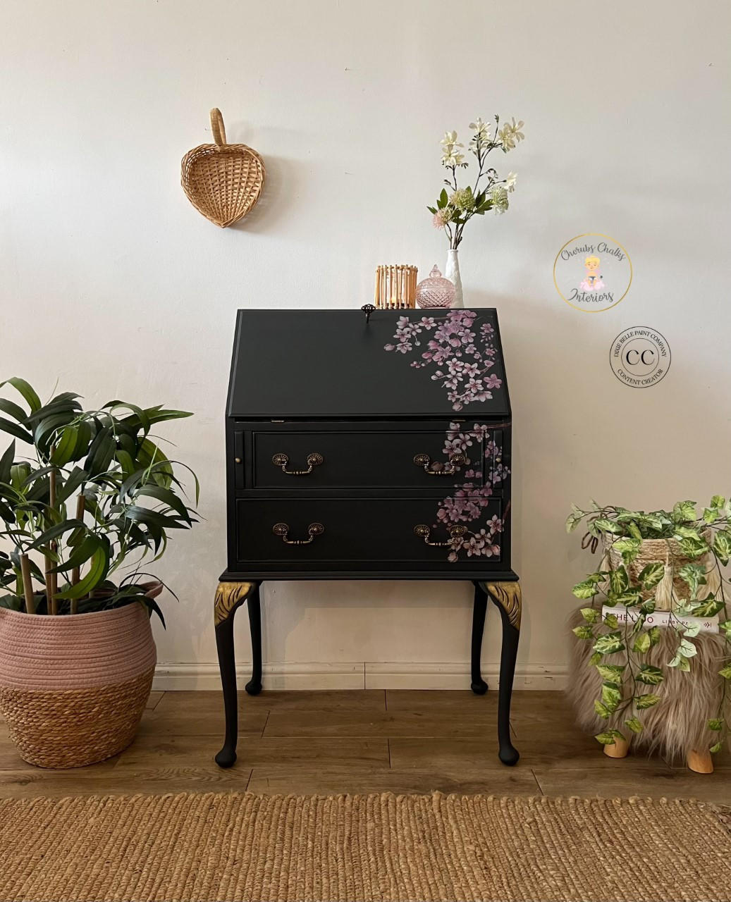 Black Sands Silk All-In-One paint is perfect for painting kitchen cabinets, bathroom cabinets and painting furniture.  Secretary desk painted in Black Sand Silk all in one paint, with gold hardware and gold accents on top of legs. Cherry blossom rub on Transfer on right side of desk. Staged with woven baskets on either side of desk on floor, right side is on top of gray fur stool. On top of desk small white vase with faux flowers and pink glass jar and wooden stick candle holder.
