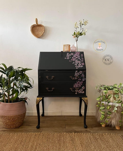 Black Sands Silk All-In-One paint is perfect for painting kitchen cabinets, bathroom cabinets and painting furniture.  Secretary desk painted in Black Sand Silk all in one paint, with gold hardware and gold accents on top of legs. Cherry blossom rub on Transfer on right side of desk. Staged with woven baskets on either side of desk on floor, right side is on top of gray fur stool. On top of desk small white vase with faux flowers and pink glass jar and wooden stick candle holder.