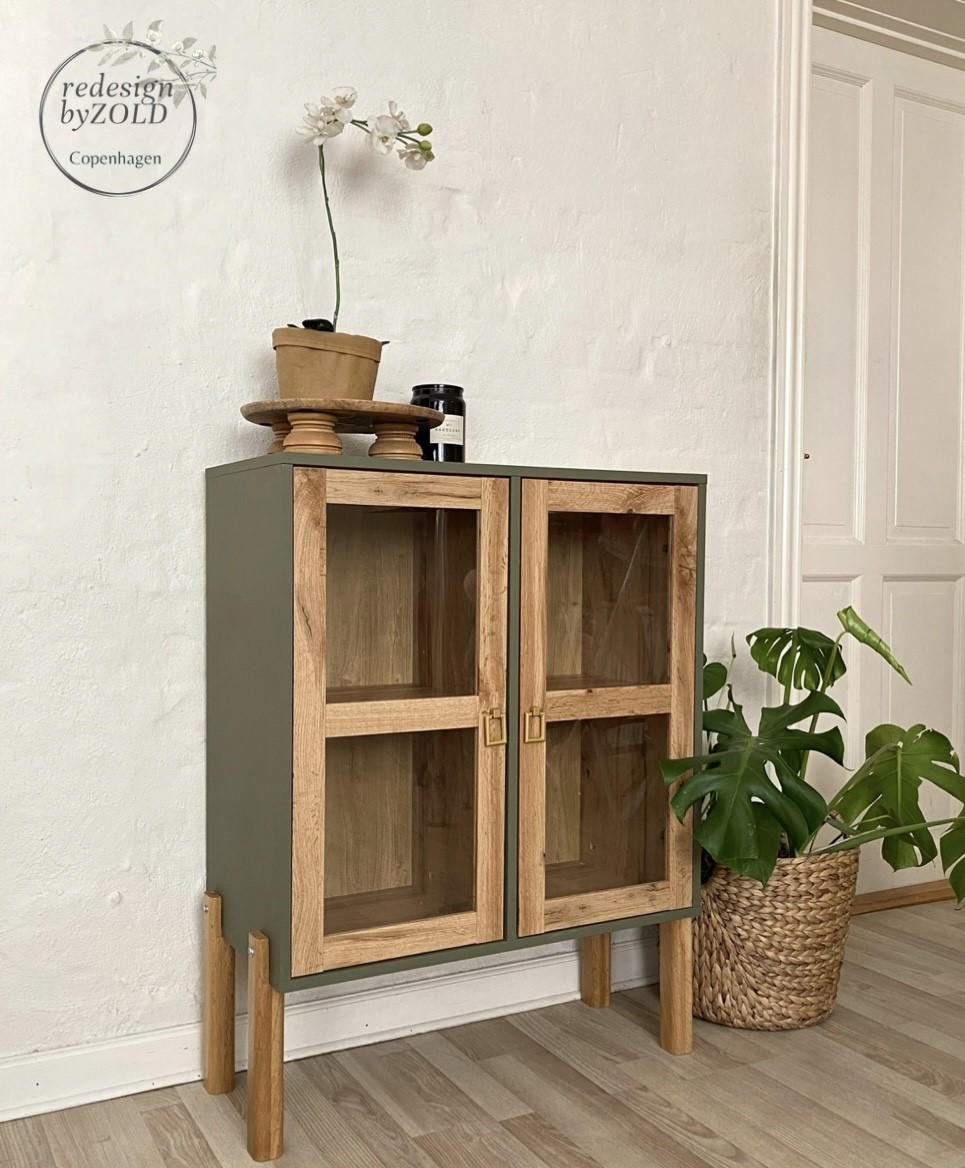 Cactus Silk All-In-One paint is perfect for painting kitchen cabinets, bathroom cabinets and painting furniture.
Cup cabinet with the body painted in Cactus Silk All-In-One paint with the legs, and door panels are left with a wood stain. Staged with a wooden tray and a brown potted orchid and a black candle is on top of the cabinet. A woven pot with a houseplant inside is on the right side.