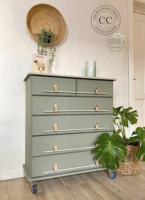 Cactus Silk All-In-One paint is perfect for painting kitchen cabinets, bathroom cabinets and painting furniture.
Chest of drawers painted in Cactus Silk All-In-One Mineral Paint® with silver metal bars as the hardware, staged with houseplants above and next to the piece on wooden floors with a white wall and a rattan wall decor.