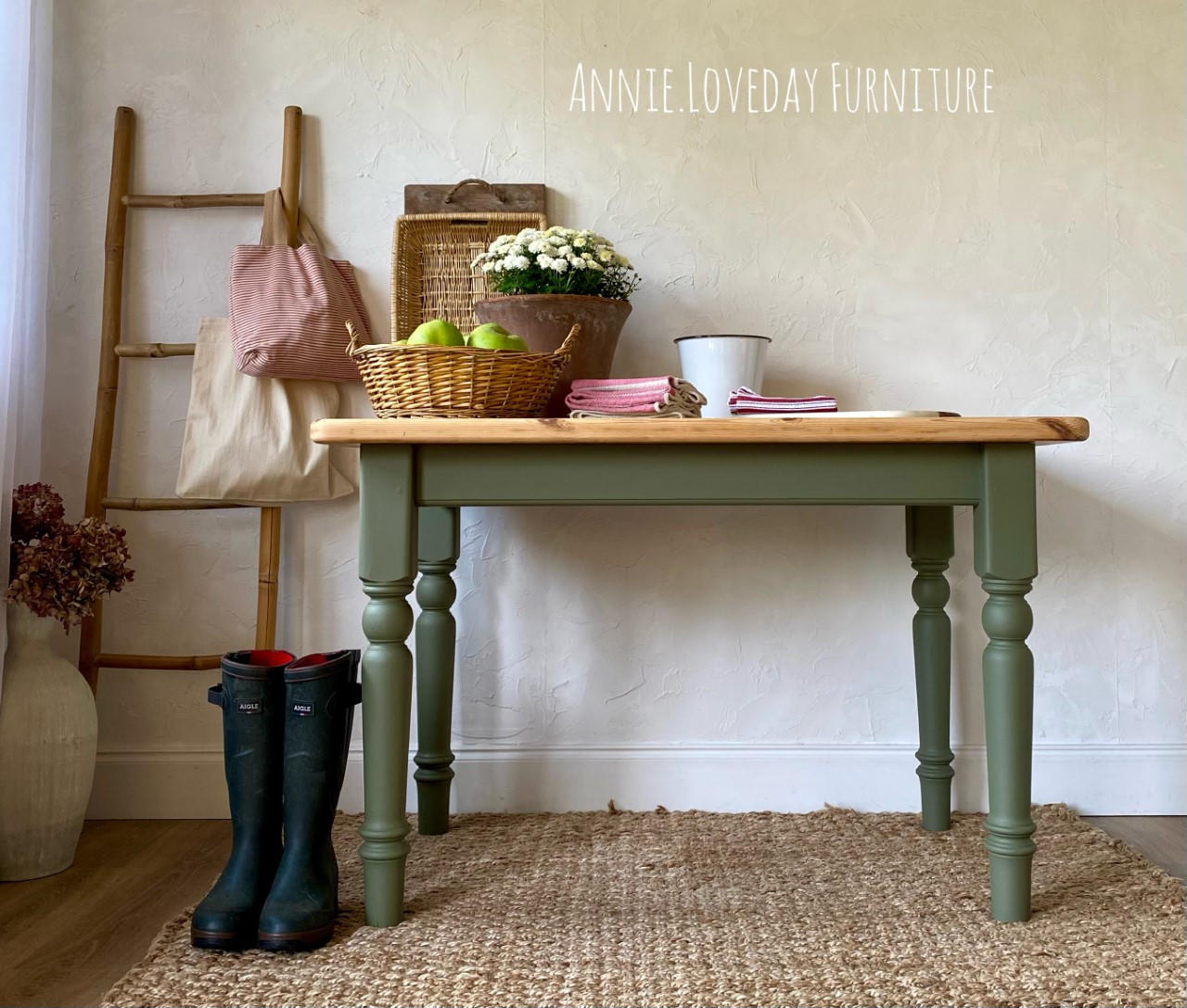 Cactus Silk All-In-One paint is perfect for painting kitchen cabinets, bathroom cabinets and painting furniture.
A table with its base and legs painted in Cactus Silk All-In-One paint with the top in a natural wood color. Staged with woven baskets and decor, books, flowers, A stick ladder with two cloth bags hanging on the ladder with a pair of black rain boots on the left side of the piece.