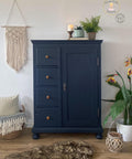 This is a tall wardrobe painted in Deep Sea Silk all-in-one mineral paint.  there are brown, round knobs to open the drawers.  On top of the wardrobe is a plant, candle stick with a white candle, a book and on top of the book is a light blue flower pot with a plant in it.  On the wall and to the left of the wardrobe is a macramé wall hanging.  Underneath that is a wicker basket with a white pillow and a blanket with tassels.  to the right of the wardrobe is a white flower pot and birds-of-paradise in it.  T